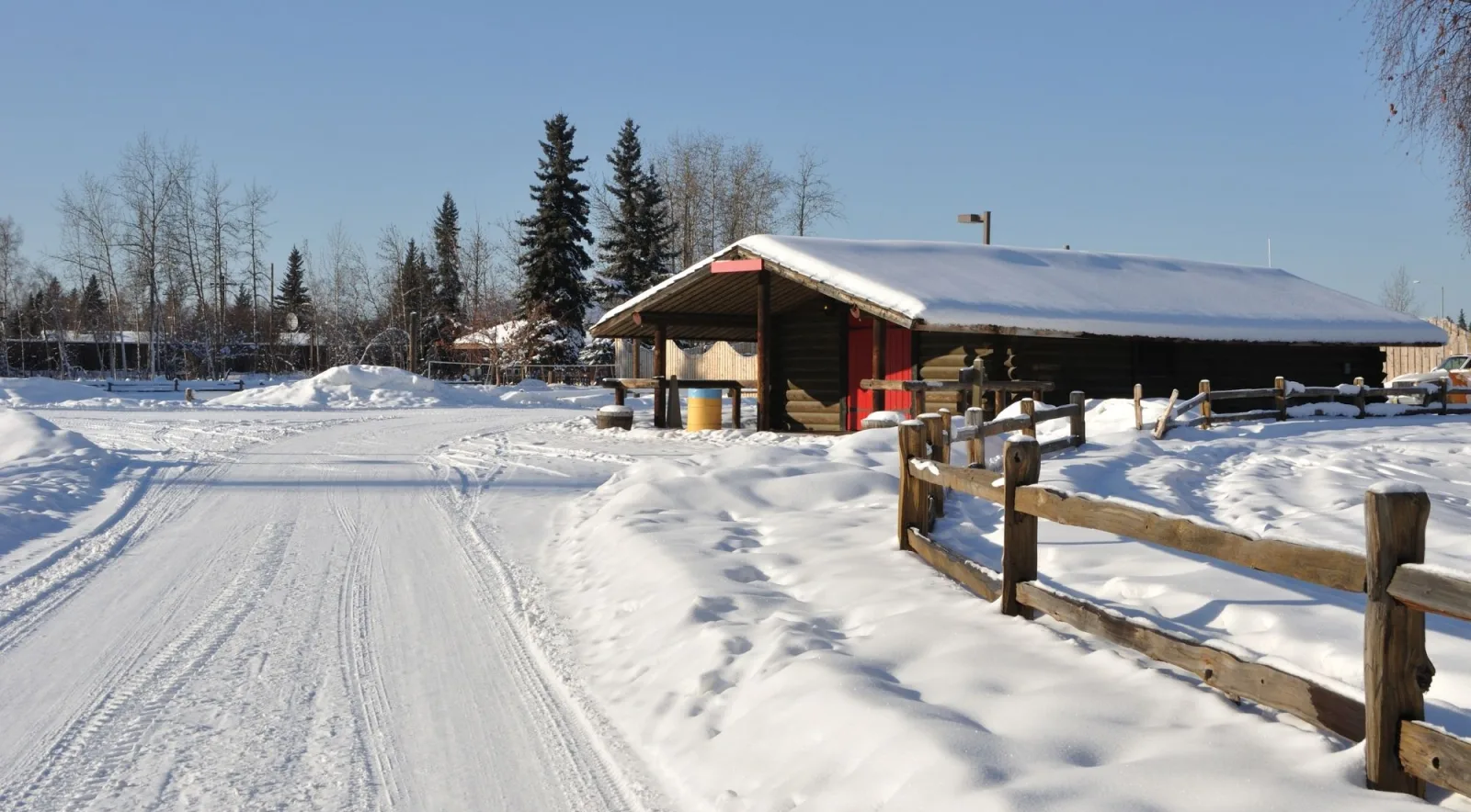 Snowy road beside a log cabin with a red door and a split-rail fence