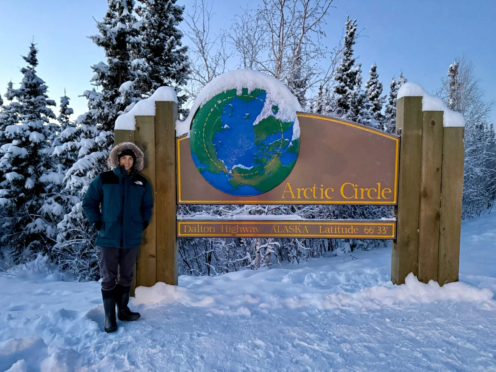 Guest in a parka beside the snow-capped Arctic Circle sign at Dalton Highway milepost 115