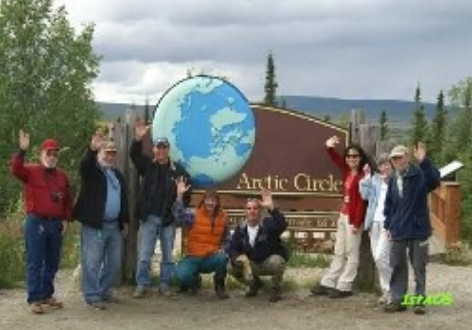 Group of 1st Alaska Tours guests waving beside the Arctic Circle sign on the Dalton Highway in summer