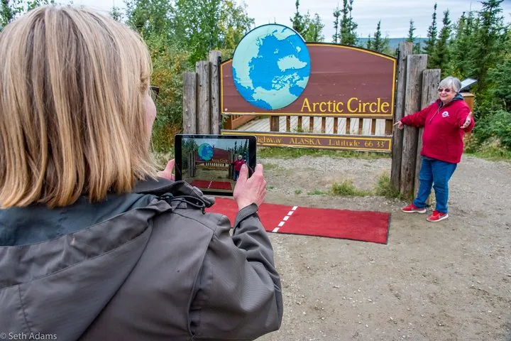 Guest taking a photo of a fellow traveler posing at the Arctic Circle sign, latitude 66 33