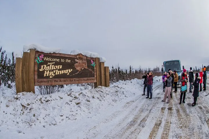 Tour group photographing the snow-topped Welcome to the James Dalton Highway sign beside the coach