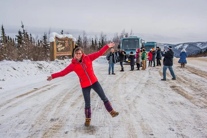 Guest posing on the icy Dalton Highway with two Northern Alaska Tour Company coaches behind