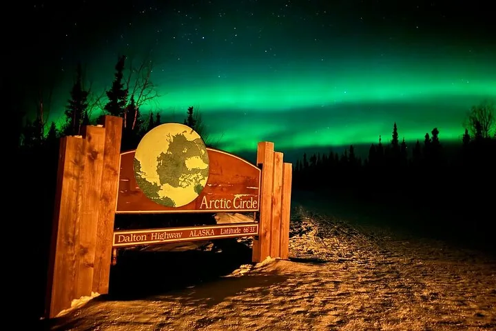 Arctic Circle sign on the Dalton Highway at night with green aurora bands filling the sky behind it