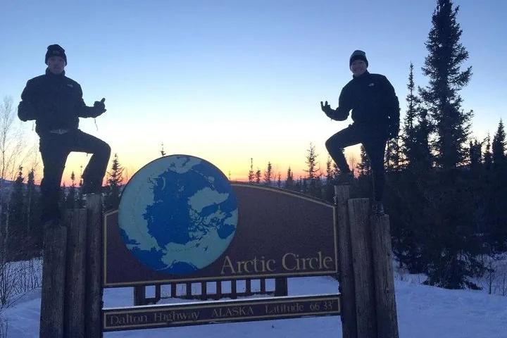 Two guests in winter gear posing on the posts of the Arctic Circle sign at sunset