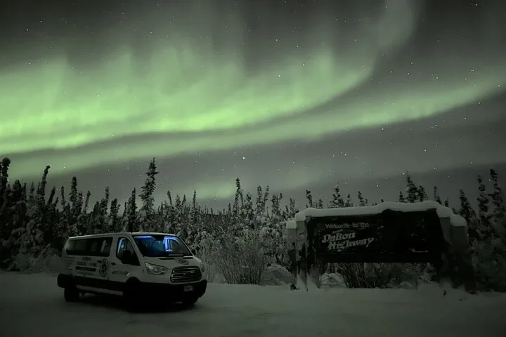 Alaska Wild Lights van parked beside the Dalton Highway welcome sign under a green aurora